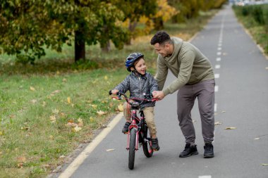 Baba oğluna parktaki bisiklet yolunda bisiklet sürmeyi öğretiyor. Baba bir bisiklet tutuyor ve oğlu üzerinde oturuyor..