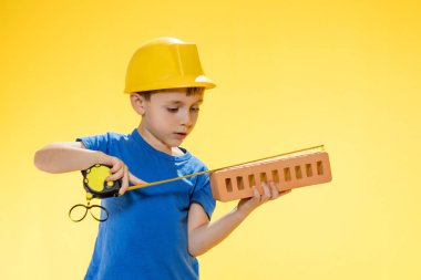 Boy in a construction helmet holds a brick in his hands and measures it with a tape measure staiding in studio on yellow background. clipart