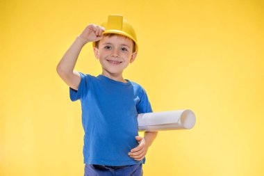 İnşaat kasklı bir çocuk elinde bir tuğla tutuyor ve onu sarı arka plandaki stüdyoda bulunan bir mezura ile ölçüyor..