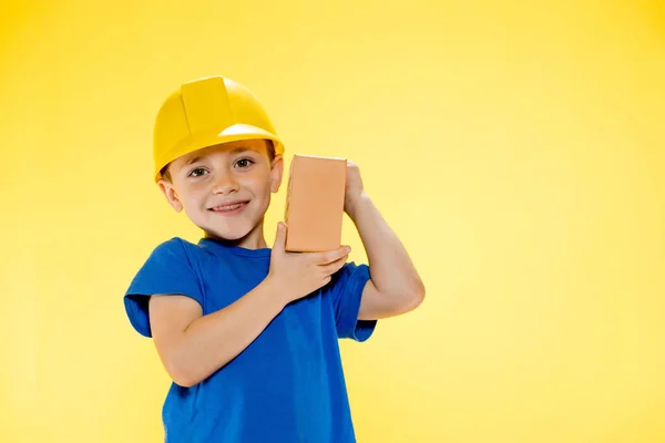 Chlapec Stavební Helmě Drží Cihlu Svých Rukou Žlutém Pozadí — Stock fotografie