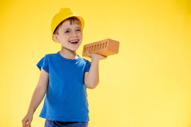 İnşaat kasklı çocuk elinde sarı arka planda bir tuğla tutuyor..