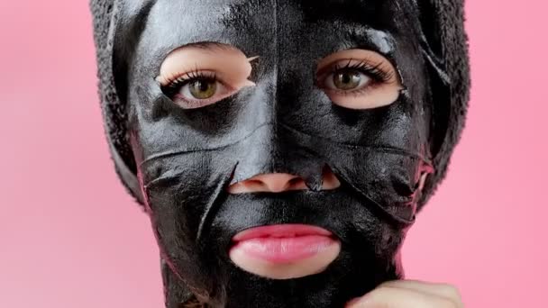 Kadın pembe arka plana siyah kozmetik yüz maskesi takıyor. Kömürle maske soyma, güzellik tedavisi, cilt bakımı, kozmetoloji — Stok video