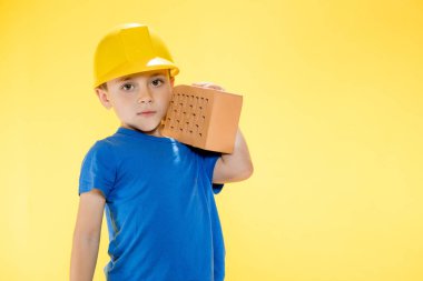 İnşaat kasklı çocuk elinde sarı arka planda bir tuğla tutuyor..