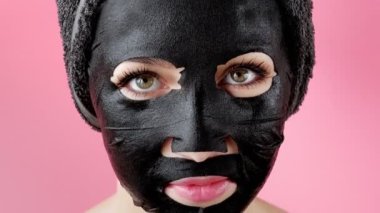 Genç bir kadın pembe arka plana siyah kozmetik yüz maskesi takıyor. Kömürle maske soyma, güzellik tedavisi, cilt bakımı, kozmetoloji.