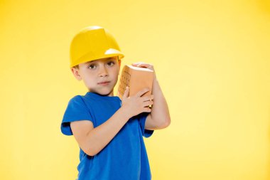 İnşaat kasklı çocuk elinde sarı arka planda bir tuğla tutuyor..