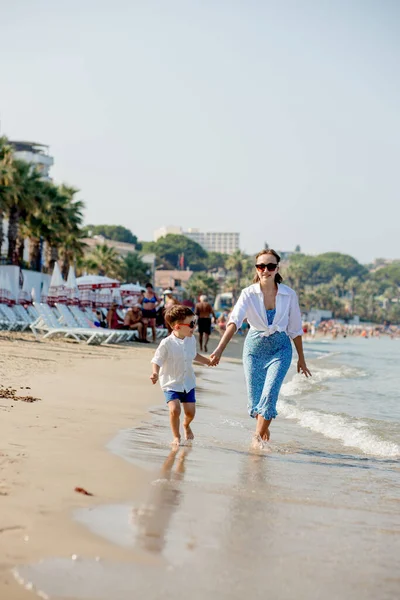 快乐的年轻妈妈和儿子在一个阳光明媚的日子在海边奔跑 在海滩上休息 幸福的家庭 — 图库照片