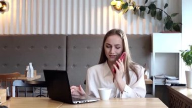 Formalite içinde genç bir iş kadını cep telefonuyla konuşur ve bir restoranda bir fincan kahveyle otururken dizüstü bilgisayar kullanır..