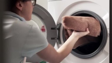 Çamaşır makinesinin önünde oturan ve çamaşır makinesini kirli havlularla dolduran genç kadın..