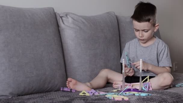 一个穿着灰色T恤的男孩坐在靠近白墙的床上 用磁力构造器制造摩天轮 创造力的发展 磁力构造函数 — 图库视频影像