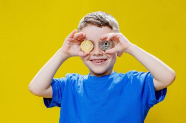 Mavi tişörtlü küçük mutlu anaokulu çocuğu sarı arka planda gözlerinin önünde ethereum ve bitcoin paralar tutuyor..