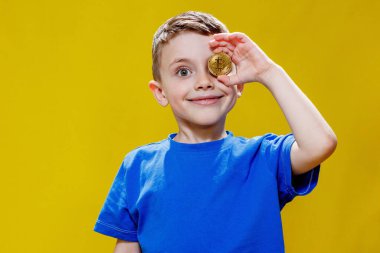 Mavi tişörtlü küçük mutlu anaokulu çocuğu sarı arka planda gözlerinin önünde ethereum ve bitcoin paralar tutuyor..