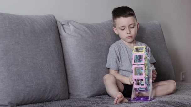 一个穿着灰色T恤的男孩坐在靠近白墙的床上，玩着他用磁力构造器设计的摩天轮。创造力的发展。磁力构造函数 — 图库视频影像