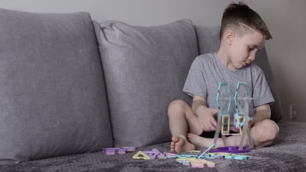 一个穿着灰色T恤的男孩坐在靠近白墙的床上，用磁力构造器制造摩天轮。创造力的发展。磁力构造函数 — 图库视频影像