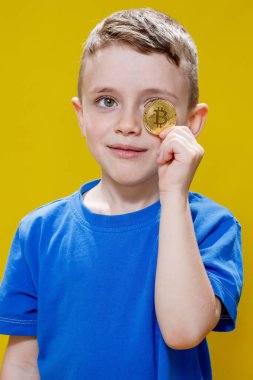 Mavi tişörtlü küçük mutlu anaokulu çocuğu sarı arka planda gözlerinin önünde Bitcoin tutuyor..