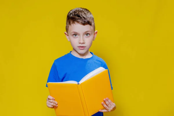 穿着蓝色T恤的学龄前男孩拿着一本黄色背景的打开的黄皮书 — 图库照片
