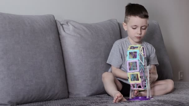 一个穿着灰色T恤的男孩坐在靠近白墙的床上 玩着他用磁力构造器设计的摩天轮 创造力的发展 磁力构造函数 — 图库视频影像