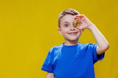 Mavi tişörtlü küçük mutlu anaokulu çocuğu sarı arka planda gözlerinin önünde Bitcoin tutuyor..