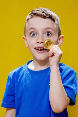 Mavi tişörtlü küçük mutlu anaokulu çocuğu sarı arka planda gözlerinin önünde Bitcoin tutuyor..