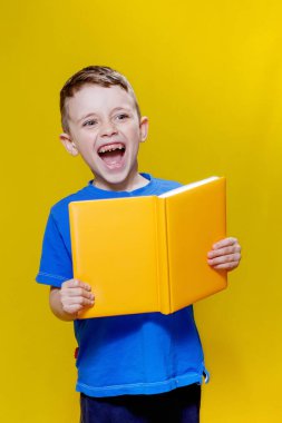 Mavi tişörtlü pozitif anaokulu çocuğu sarı arka planda açık sarı bir defter tutuyor..