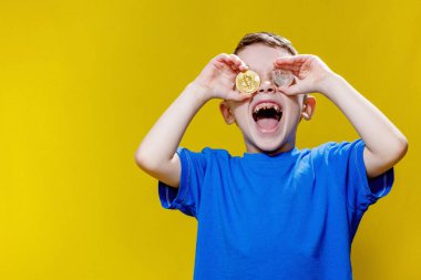 Mavi tişörtlü küçük mutlu anaokulu çocuğu sarı arka planda gözlerinin önünde ethereum ve bitcoin paralar tutuyor..