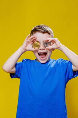 Mavi tişörtlü küçük mutlu anaokulu çocuğu sarı arka planda gözlerinin önünde ethereum ve bitcoin paralar tutuyor..