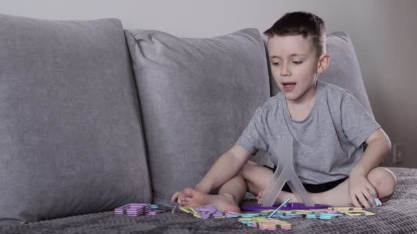 一个穿着灰色T恤的男孩坐在靠近白墙的床上 用磁力构造器制造摩天轮 创造力的发展 磁力构造函数 — 图库视频影像
