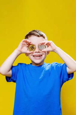 Mavi tişörtlü küçük mutlu anaokulu çocuğu sarı arka planda gözlerinin önünde ethereum ve bitcoin paralar tutuyor..