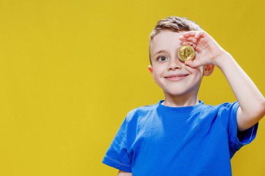 Mavi tişörtlü küçük mutlu anaokulu çocuğu sarı arka planda gözlerinin önünde Bitcoin tutuyor..