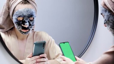 Aynanın önündeki kadın yüz derisine dikkat etmek için nemlendirici bir maske takar ve cep telefonuna bakar..