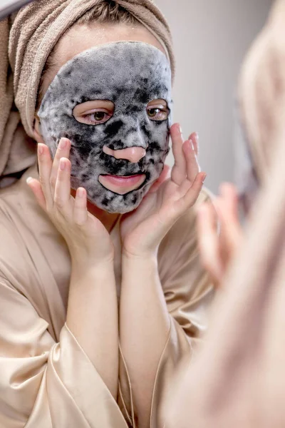Young happy woman in towel making face mask and looking in mirror in stylish bathroom. The woman uses a mask to cleanse the skin. Skin care.