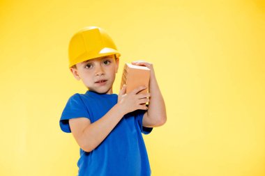 İnşaat kasklı çocuk elinde sarı arka planda bir tuğla tutuyor..