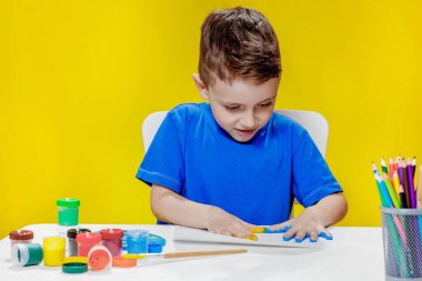 Küçük bir anaokulu öğrencisi masada oturuyor, avuç içlerini bir kağıt parçasına çiziyor. Renkli el izleri. Anaokulu geliştirme.