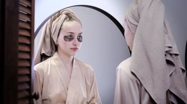 Şişlik için göz bantları, kırışıklıklar, siyah halkalar. Göz bantları. Krem rengi elbiseli tatmin olmuş kadın banyodaki aynaya bakan gözlere kahverengi bantlar yapıştırıyor..