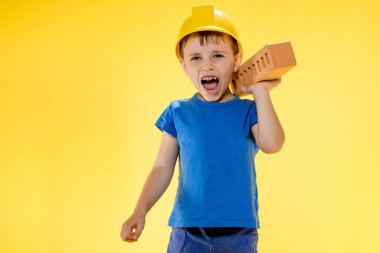 İnşaat kasklı çocuk elinde sarı arka planda bir tuğla tutuyor..