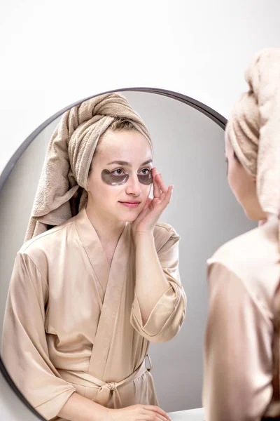 Eye patches for swelling, wrinkles, dark circles. Eye patches. Satisfied woman in cream gown applies brown patches to eyes in bathroom looking in mirror.