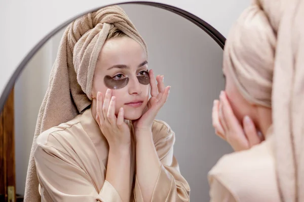 Eye patches for swelling, wrinkles, dark circles. Eye patches. Satisfied woman in cream gown applies brown patches to eyes in bathroom looking in mirror.