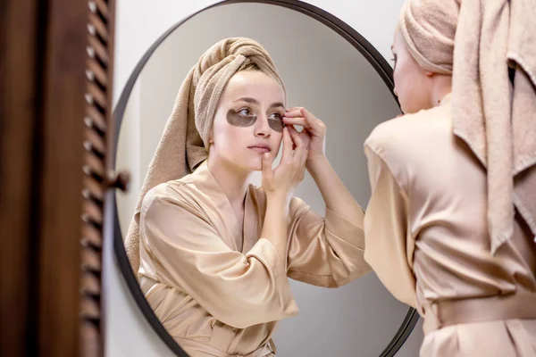 Bej rengi havlu ve bornozlu genç bir kadın, göz altı yamalı deriyle ilgileniyor.