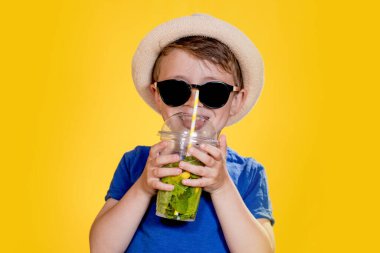 Yazlık kıyafetli çocuk güneş gözlüğü takıyor ve Mojito kokteyli içerken eğleniyor. Sarı arkaplanda poz