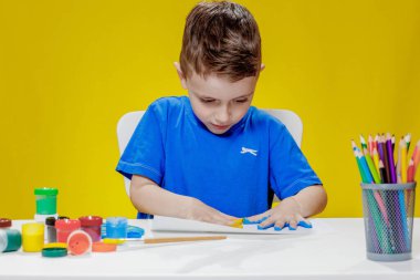 Küçük bir anaokulu öğrencisi masada oturuyor, avuç içlerini bir kağıt parçasına çiziyor. Renkli el izleri. Anaokulu geliştirme.