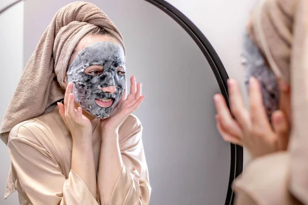 Young happy woman in towel making face mask and looking in mirror in stylish bathroom. The woman uses a mask to cleanse the skin. Skin care.