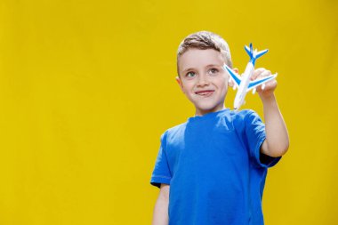 Mavi tişörtlü küçük bir çocuk oyuncak beyaz bir uçak tutuyor ve sarı arka planda oynuyor. Uzayı kopyala Seyahat kavramı.