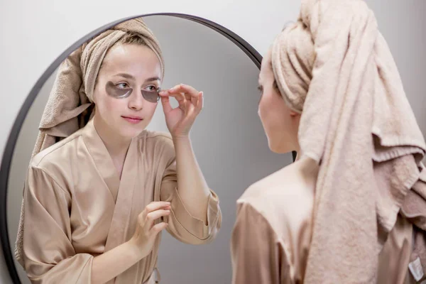 Eye patches for swelling, wrinkles, dark circles. Eye patches. Satisfied woman in cream gown applies brown patches to eyes in bathroom looking in mirror.