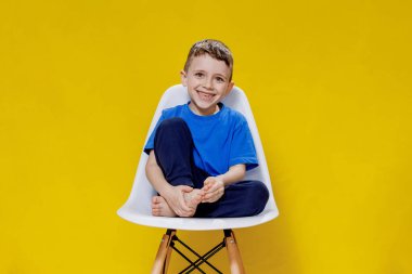 Mavi tişörtlü ve pantolonlu neşeli küçük çocuk sarı arka plan stüdyosunda beyaz bir sandalyede oturuyor..
