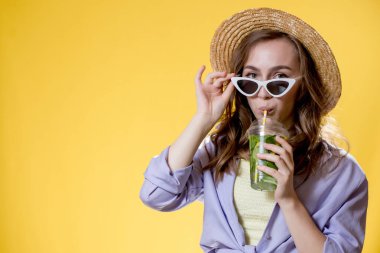 Beyaz güneş gözlüğü takan ve Mojito kokteyli içerken eğlenen yaz kıyafetli bir kadın. Sarı arkaplanda poz.