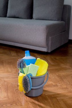 Bucket with cleaning items in the house, standing on the floor, on the background of the sofa. Housework. Cleaning.