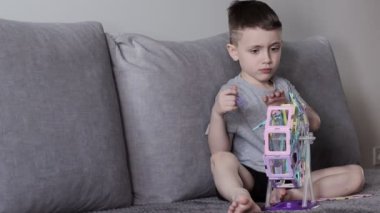 Gri tişörtlü bir çocuk beyaz bir duvarın yakınındaki bir yatakta oturur ve manyetik bir yapıcıyla tasarladığı dönme dolapla oynar. Yaratıcılık gelişimi. Manyetik yapıcı.