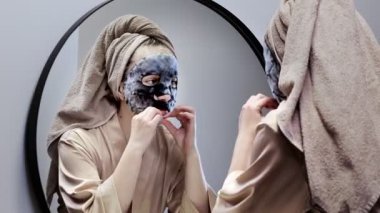 Havlu giymiş, yüz maskesi yapan ve şık banyoda aynaya bakan mutlu genç bir kadın. Kadın deriyi temizlemek için maske kullanıyor. Cilt bakımı