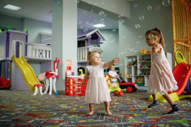 Two little sisters playing with soap bubbles, dancing, celebrating birthday, being in a good mood, in the playroom. Childhood concept.