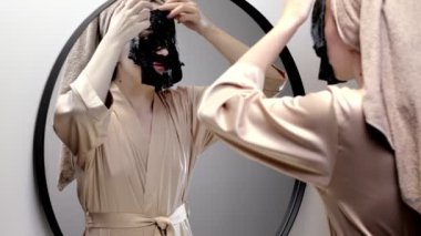 Havlu giymiş, yüz maskesi yapan ve şık banyoda aynaya bakan mutlu genç bir kadın. Kadın deriyi temizlemek için maske kullanıyor. Cilt bakımı.