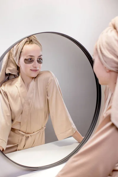 Eye patches for swelling, wrinkles, dark circles. Eye patches. Satisfied woman in cream gown applies brown patches to eyes in bathroom looking in mirror.